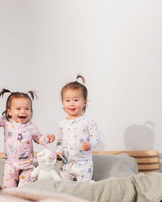 Two children in pajamas standing on a bed with a plush toy and a stuffed animal.